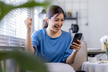 Young Happy Asian woman smiling holding Phone with fist hand and excited for success on sofa at...