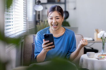 Young Happy Asian woman smiling holding Phone with fist hand and excited for success on sofa at...