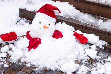 Unhappy snowman in mittens, red scarf and cap is melting  outdoors in sunlight on wet pavement Approaching spring, warm winter, climate change concept  