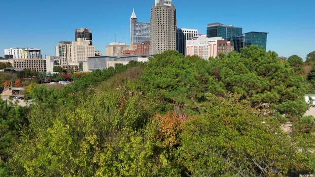 Aerial reveal of Raleigh skyline from behind green trees during autumn day. Raleigh, North Carolina cityscape establishing shot.