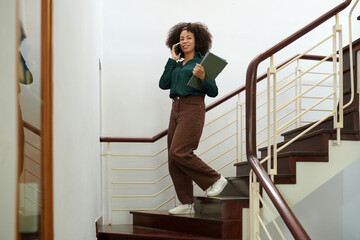 Smiling woman with clipboards talking on phone when walking down stairs