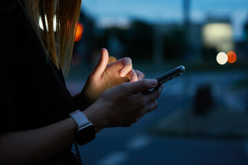 Close up shot of woman using smartphone on city street with bokeh lights at night. Mobile phone in...
