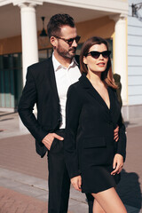 Beautiful fashion woman and her handsome elegant boyfriend in black suit. Sexy brunette model in jacket. Fashionable confident couple posing in street. Brutal man and female outdoors. In sunglasses