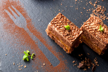 Chocolate cake cut into square pieces with nut cream. Dessert on black surface with fork reflection