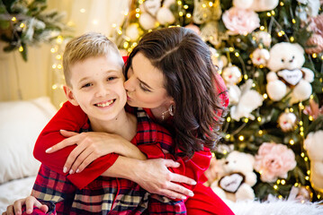 Fototapeta na wymiar smiling happy mother and son wearing red pajamas posing and hugging near christmas tree in decorated living room