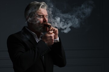 Bearded man lighting cigar on dark grey background. Space for text