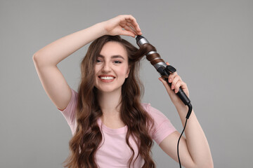 Beautiful young woman using curling hair iron on grey background