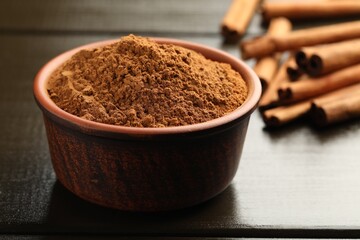 Bowl of cinnamon powder and sticks on wooden table, closeup
