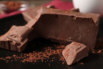 Pieces of tasty milk chocolate and cocoa powder on black plate, closeup