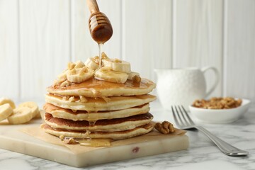 Pouring honey from dipper onto delicious pancakes with bananas and walnuts at white marble table,...