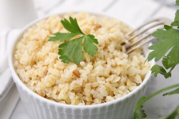 Cooked bulgur with parsley in bowl on white table, closeup