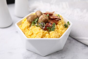 Cooked cornmeal with bacon, mushrooms and microgreens in bowl on white marble table, closeup