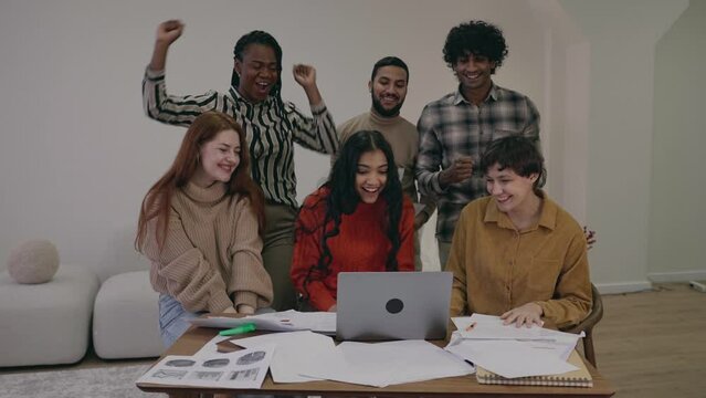 Happy multiracial multiethnic excited students doing research project, looking at laptop, feeling euphoria success. The work or study is finished. The start of holidays vacation. People raising hands