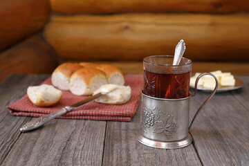 A glass of hot tea in a metal cup holder next to delicious buns on a red textile and a metal plate with butter are on a wooden background.