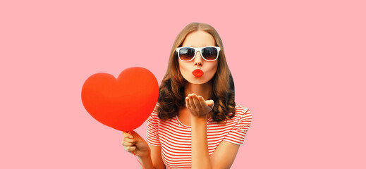 Portrait of happy young woman with red heart shaped balloon blows her lips sends air kiss wearing sunglasses on pink studio background