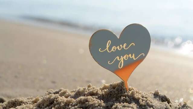 Plastic stick shape of heart with words Love you in sand beach of sea shore on background sea waves on sunny summer day close-up. Concept love relationship romance amour celebration St Valentines Day