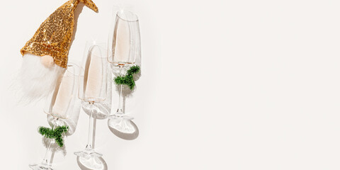Minimal New Year flat lay with white wine in champagne glasses, Christmas green tinsel, golden gnome toy for bottle decor. Holiday aesthetic still life banner, top view wineglass, sunlight, star