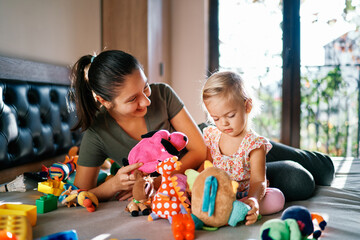 Smiling mom playing with little girl soft toys while sitting on bed in room