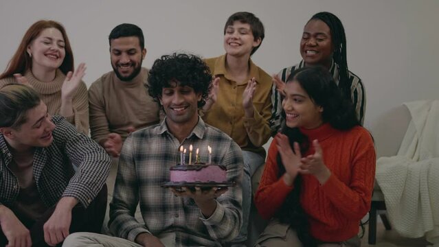 Happy indian male having birthday party, blowing candles celebrating with multiracial friends at home with lights, special event with friends
