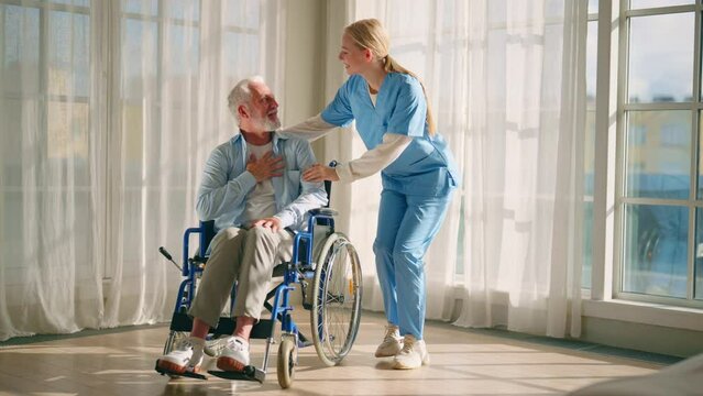 Social worker helps man in wheelchair. Female social welfare physical therapist looks after senior male, talking, providing medical care at home. Living with legs paralysis, special needs people.