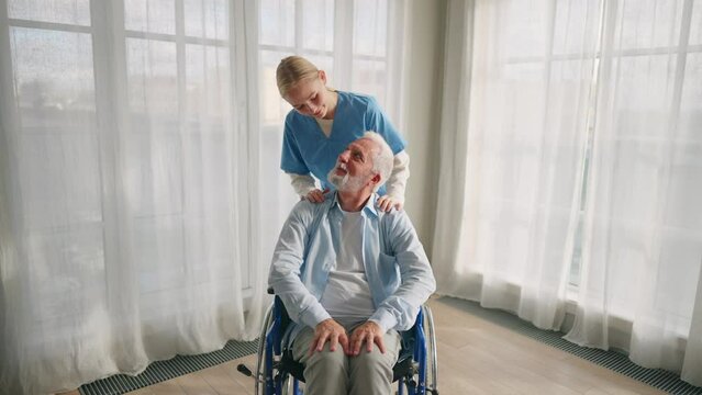 Nurse massaging neck to paralyzed man. Senior male in wheelchair enjoying massage at home. Social medical worker provides care support help. Living with legs paralysis, special needs people concept.
