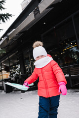 Kid playing in snowy, having fun with snow on street. Funny little girl in red winter clothes walks during snowfall. Outdoors winter activities. Cute child wearing a warm hat catching snowflakes.