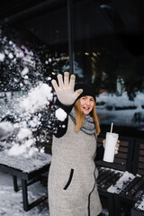 Winter smiling woman in snow covered street. Young female drinking coffee, tasty cocoa, or hot tea outdoors. Girl rest and walk in winter park. Closeup. Beautiful fashionable girl during snowfall.
