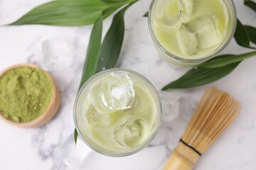 Glasses of tasty iced matcha latte, bamboo whisk and leaves on white marble table, flat lay