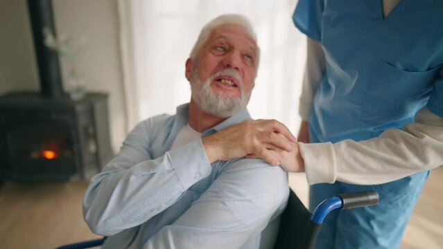 Old man in wheelchair talks with nurse. Senior paralyzed male thanks social worker woman for help support at home. Caregiver holds hand on patient shoulder stands near. Legs paralysis, special needs.