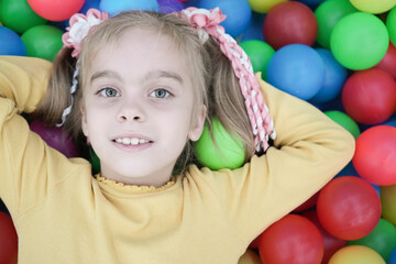 Fototapeta na wymiar Portrait of a beautiful blond girl with two ponytails against a background of multi-colored bright balls. Close-up. Beautiful happy girl in the playroom.
