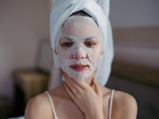 Young woman lying on bed with moisturizing beauty face mask and white towel on her head after shower, lifestyle home skincare, smile.