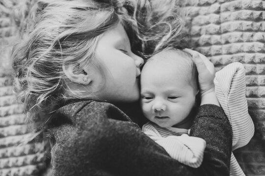 Baby top view closeup. Girl tenderly hugging and kissing newborn while lying on bed at home together. Sibling relationship in family. First meeting baby and toddler older sister. Black and white photo