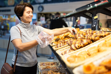 Mature woman chooses hot fresh sweet buns at supermarket showcase