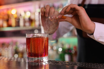 Bartender spraying orange peel onto fresh alcoholic cocktail at counter in bar, closeup