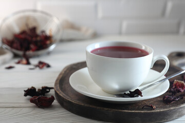 Delicious hibiscus tea in cup and dry roselle petals on white wooden table, closeup. Space for text