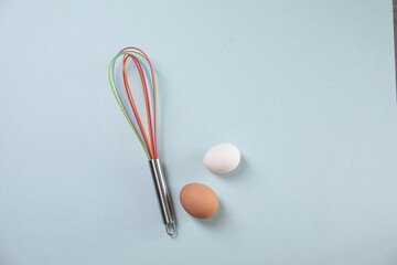 Colorful whisk and raw eggs on light blue background, flat lay