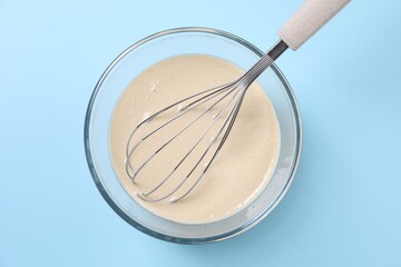 Dough and whisk in glass bowl on light blue background, top view