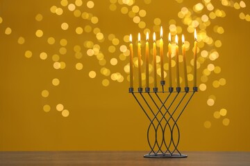 Hanukkah celebration. Menorah with burning candles on table against yellow background with blurred lights, space for text