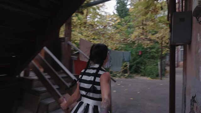 A girl is running into the tunnel and then into local houses area, dressed in a Halloween costume with dark makeup and holding a book. The scene is in slow motion during the daytime.