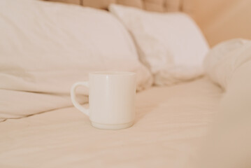 Fototapeta na wymiar White coffee cup on the bed in sunny morning bedroom, mock up.