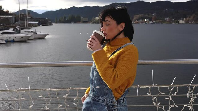 young adult woman walks along the port while drinking coffee and looking at her mobile phone