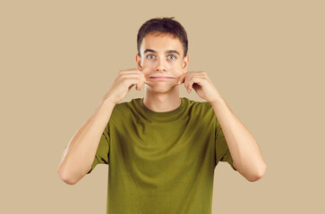 Close up studio portrait of young grimacing boy student wearing casual green t-shirt having fun pulling back his cheeks isolated on a beige background. People emotions, facial expression concept. - obrazy, fototapety, plakaty