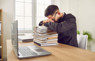 Tired stressed young business man accountant in suit working at the desk putting head on a pile of...