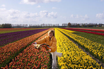 A young woman in a yellow jacket catches a kerchief torn off by the wind in a field with colorful...