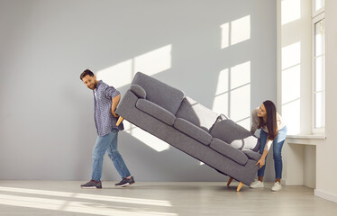 Happy young couple carrying sofa to their new home. Cheerful husband and wife placing sofa in empty...