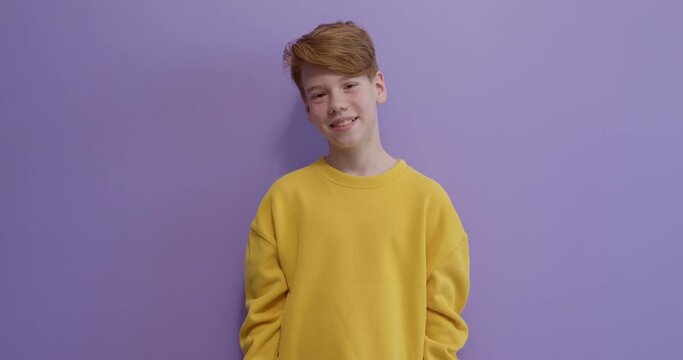 nice caucasian redhead boy in yellow shirt isolated on purple background. People lifestyle concept. Mock up copy space. Waving greeting with hand as notices someone, smiling