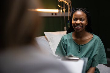 African lady in the hospital room seated on the bed, doctor across, noting the patient's medical...