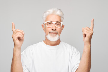 Gray-haired mature man 60 years old in white T-shirt pointing up finger, demonstrating empty space for product or text isolated on white studio background.