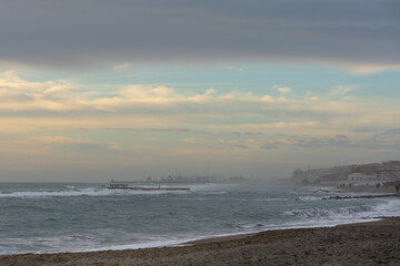 Strong gale force winds on the Mediterranean coast, a view of the coastline, buildings and people walking in the distance. Gray-white clouds in the sky with blue gaps