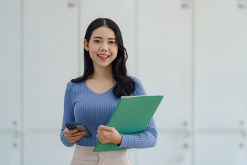 Fototapeta premium Young beautiful Asian business woman holding a folder and a smart phone while standing at the workplace.